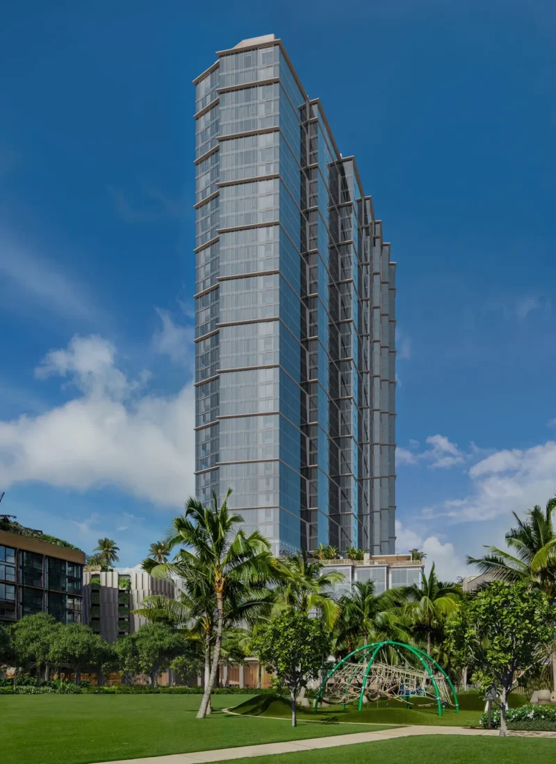 A modern high-rise building with glass windows stands behind palm trees and a grassy park under a blue sky with scattered clouds.